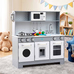 A children's toy kitchen playset with a gray and white cabinet base, faux stovetop, microwave, sink, and hanging utensils, shown in a styled indoor room with a teddy bear and shelf in the background.