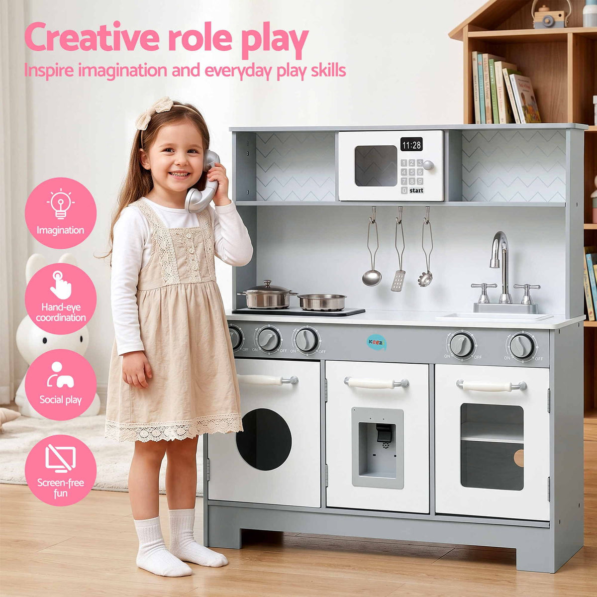  young girl stands beside a children's wooden play kitchen set in a bright indoor room. The kitchen has white and gray cabinets, toy appliances including a microwave and sink, hanging utensils, and a phone on the counter.

Details