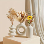 Three white vases with dried plants and flowers on a light surface.