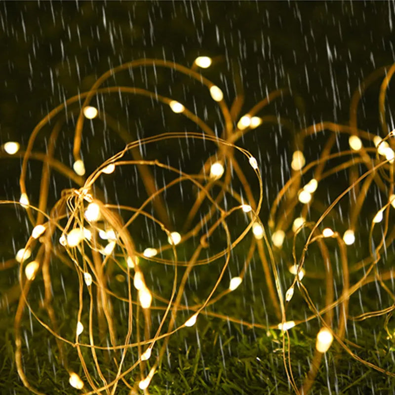 Warm white LED fairy string lights on thin copper wire, draped in loops over green grass in the rain at night.