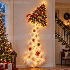 A tall, narrow Christmas tree decoration with a star-topped green top, warm white string lights, red ribbon, and small gift and snowflake ornaments, displayed indoors against a cream wall in a decorated holiday home.