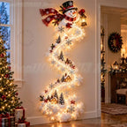 A festive Christmas wall decoration shaped like a spiral staircase, featuring a smiling snowman at the top, warm white string lights, white snowflakes, small evergreen trees, cozy house motifs, and red berry accents, displayed in a decorated home interior next to a Christmas tree.