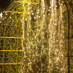 Warm white LED curtain string lights draped over a garden trellis at night, creating a glowing golden canopy against a dark background.