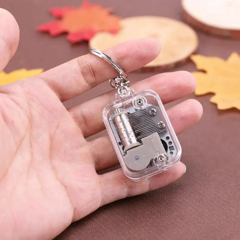 Hand holding a transparent music box keychain with mechanical components on a brown background.