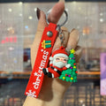 A hand holds a red Christmas-themed keychain with a long strap reading 'Christmas' in white, a small Santa Claus figure, and a colorful Christmas tree charm against a blurred indoor background.