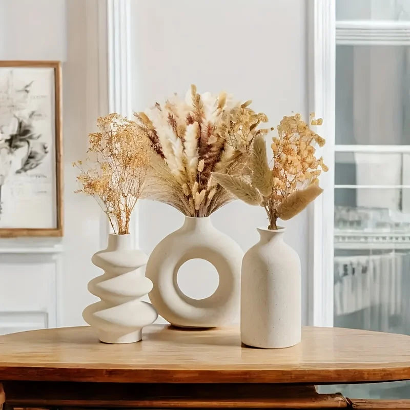 Three white ceramic vases with dried plants on a wooden surface in a room.