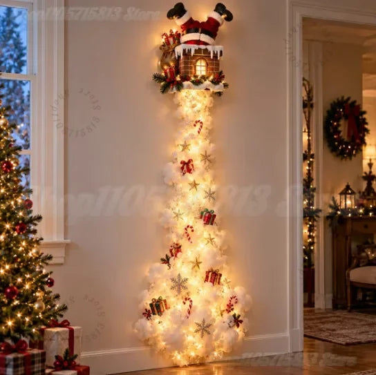 A festive Christmas tree-shaped light fixture shaped like a Santa Claus house at the top, with glowing warm white lights cascading down in a tree shape decorated with snowflake ornaments, candy canes, gifts, and red bows, set against a beige wall in a decorated holiday room.