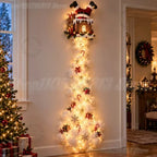 A festive Christmas tree-shaped light fixture shaped like a Santa Claus house at the top, with glowing warm white lights cascading down in a tree shape decorated with snowflake ornaments, candy canes, gifts, and red bows, set against a beige wall in a decorated holiday room.