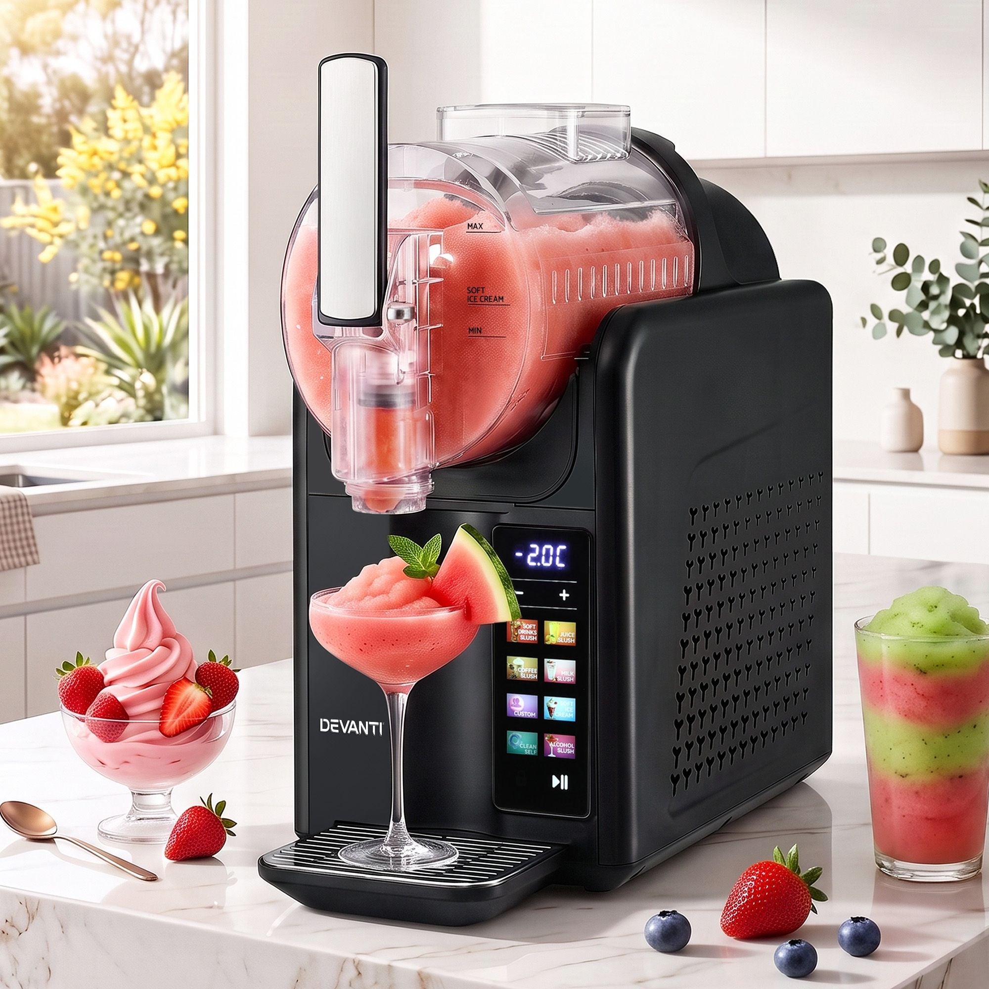 A Devanti frozen dessert machine in black sits on a kitchen counter, blending pink fruit into a slushy and dispensing it into a glass. The kitchen background is bright and clean, with additional fruit and dessert glasses styled around the coun