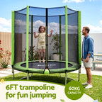 A round outdoor trampoline with a green safety net enclosure is shown in a backyard setting. A child is jumping on the trampoline while a man stands beside it.