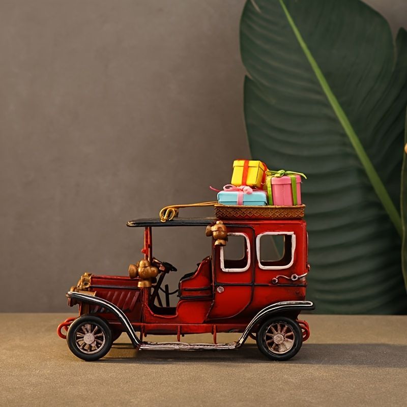 A vintage-style red toy truck or carriage with black trim, small decorative figures, and colorful gift boxes on top, photographed on a tabletop with a gray wall and large green leaf in the background.