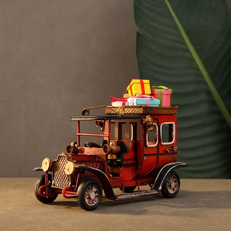 A decorative vintage red carriage-style toy or model car with a roof rack carrying colorful gift boxes, photographed on a tabletop against a gray wall with a large green leaf backdrop.