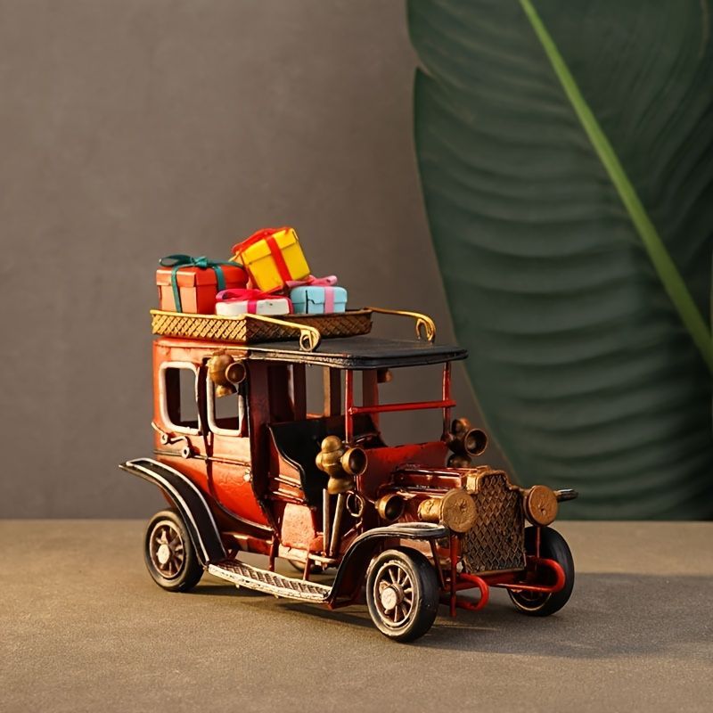A vintage-style metal model car in reddish-brown and black, carrying a stack of colorful gift boxes on its roof, photographed against a neutral gray backdrop with a large green leaf prop.