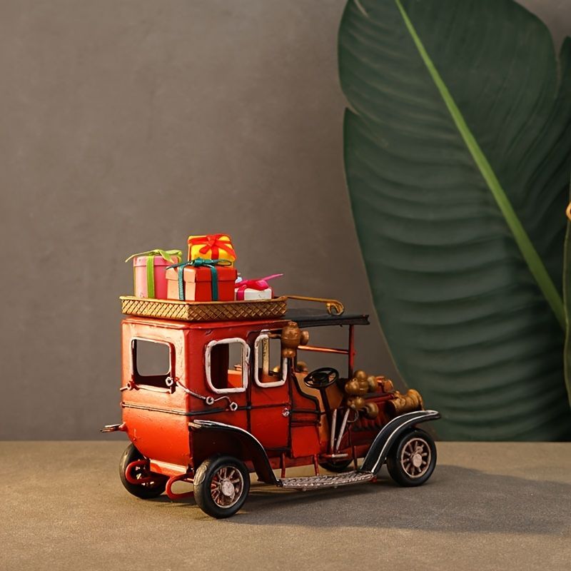 A decorative miniature red vintage jeep-style toy truck carrying colorful gift boxes, displayed on a tabletop with a large green leaf prop against a gray background.