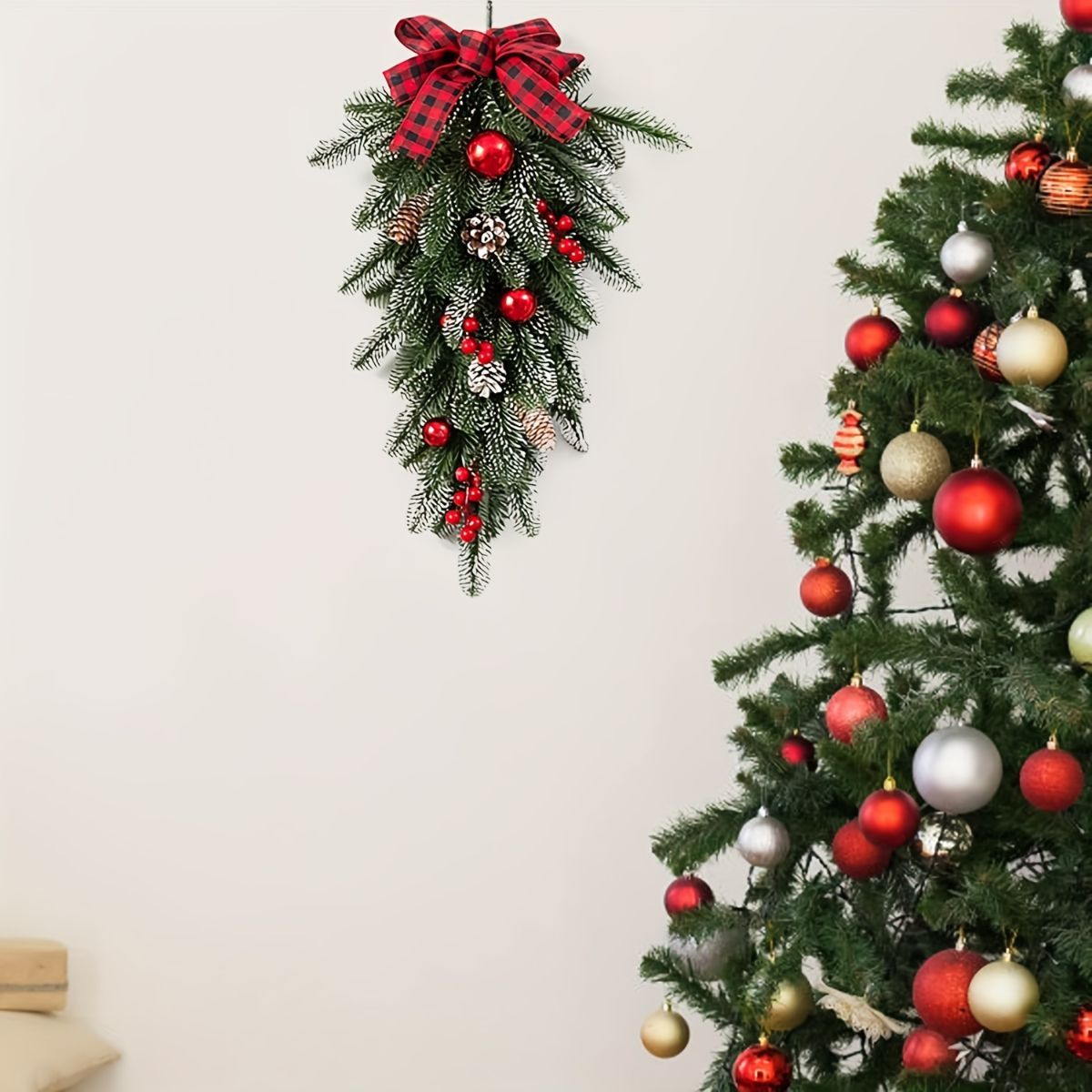 A hanging Christmas wreath decoration with green pine branches, red ornaments, pinecones, and a red plaid bow, shown against a plain light wall next to a decorated Christmas tree.