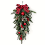 A hanging Christmas wreath decoration made of green pine branches with white frost, a red and black buffalo plaid bow at the top, red ornaments, pinecones, and red berry clusters, shown on a plain white background.