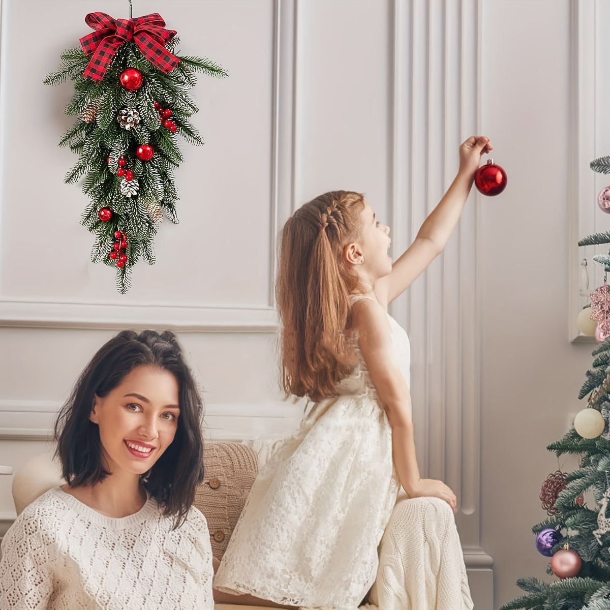 A woman and a young girl decorate a Christmas tree in a bright living room. The woman sits on a couch smiling, while the girl in a white dress reaches for a red ornament.
