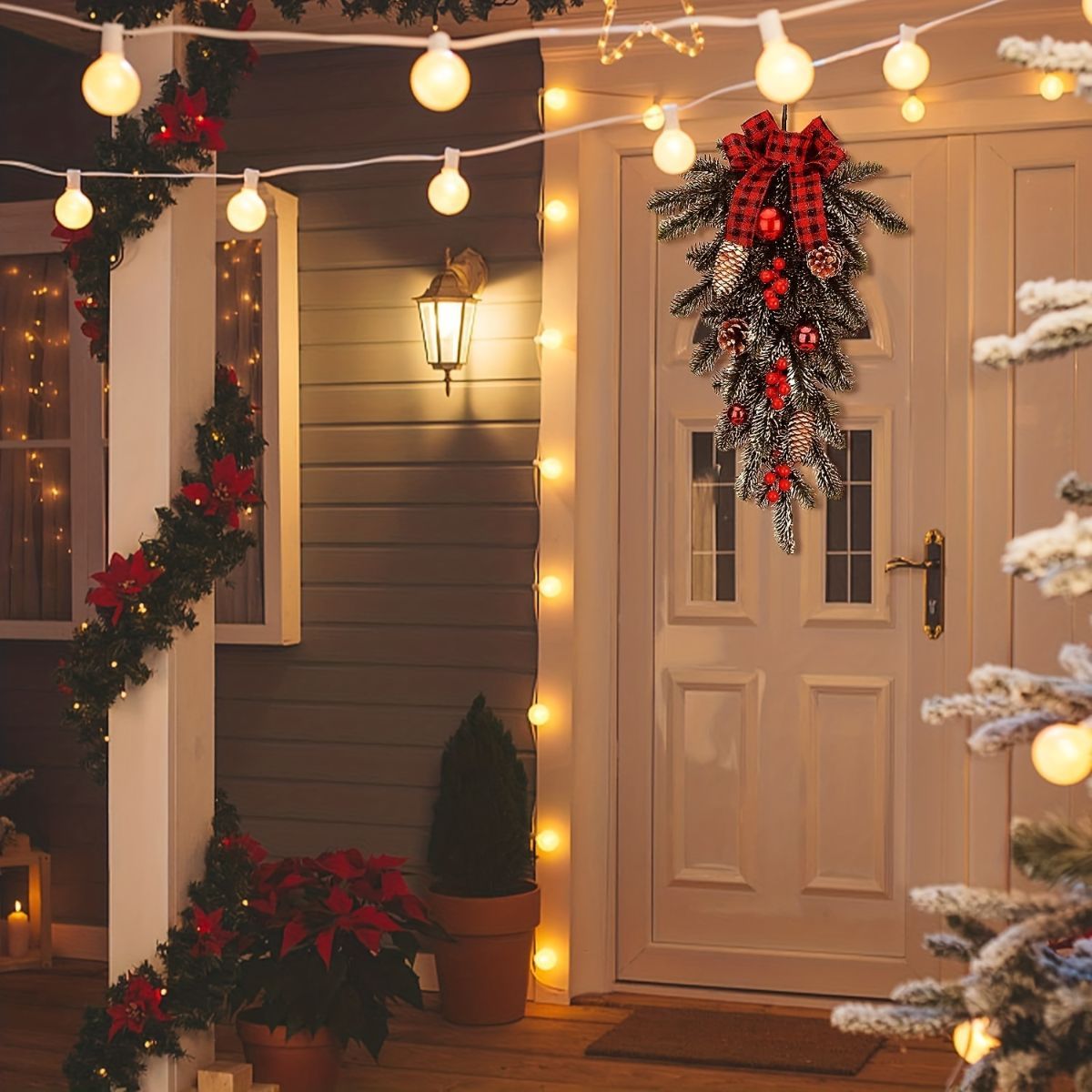 A festive Christmas wreath with a red buffalo plaid bow, silver pinecones, and red berries hangs on a white front door. The porch is decorated with warm white globe string lights, a matching green garland draped over a white column, potted poinsettias, and a small Christmas tree in the foreground.