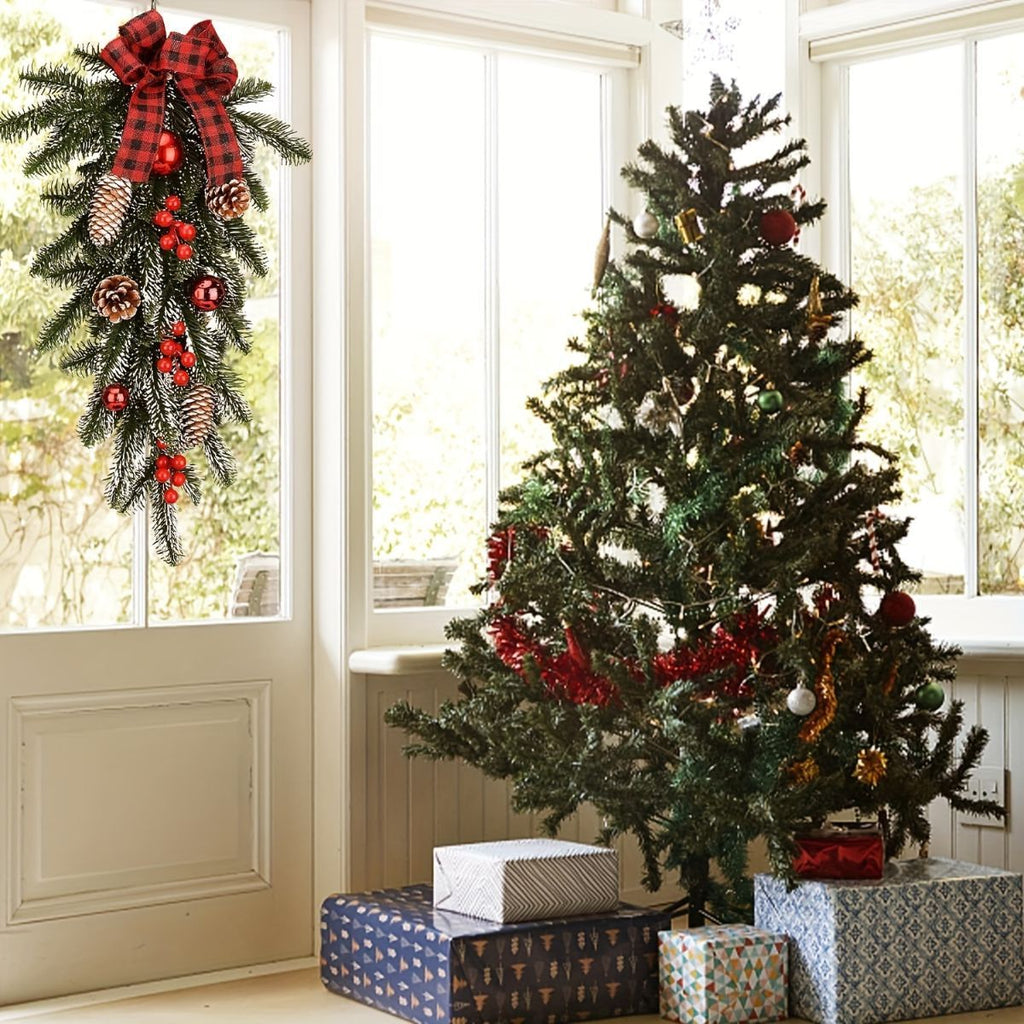 A decorated artificial Christmas tree stands indoors near large windows, with wrapped gift boxes underneath and a red plaid wreath hanging on the window beside it.