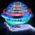 A glowing spherical grid toy floating above an open hand against a starry dark background. The sphere emits bright blue, red, green, and orange light trails, with a small miniature house visible inside the cage-like structure.