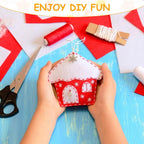Red and white house-shaped craft item held by hands with sewing materials on a blue surface.