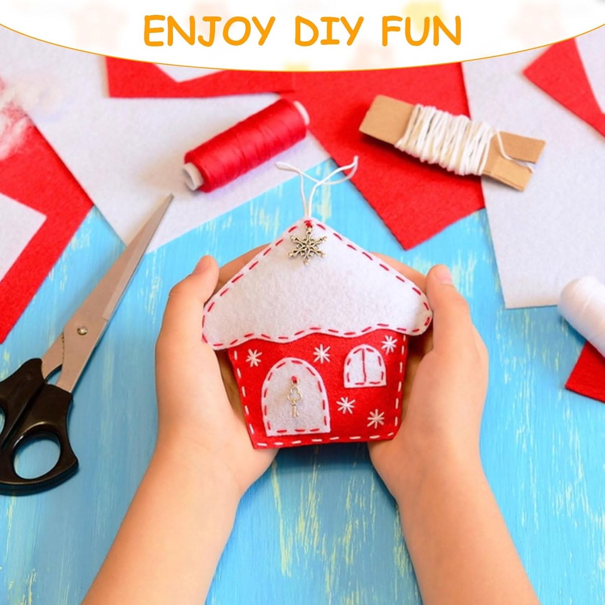 Red and white house-shaped craft item held by hands with sewing materials on a blue surface.