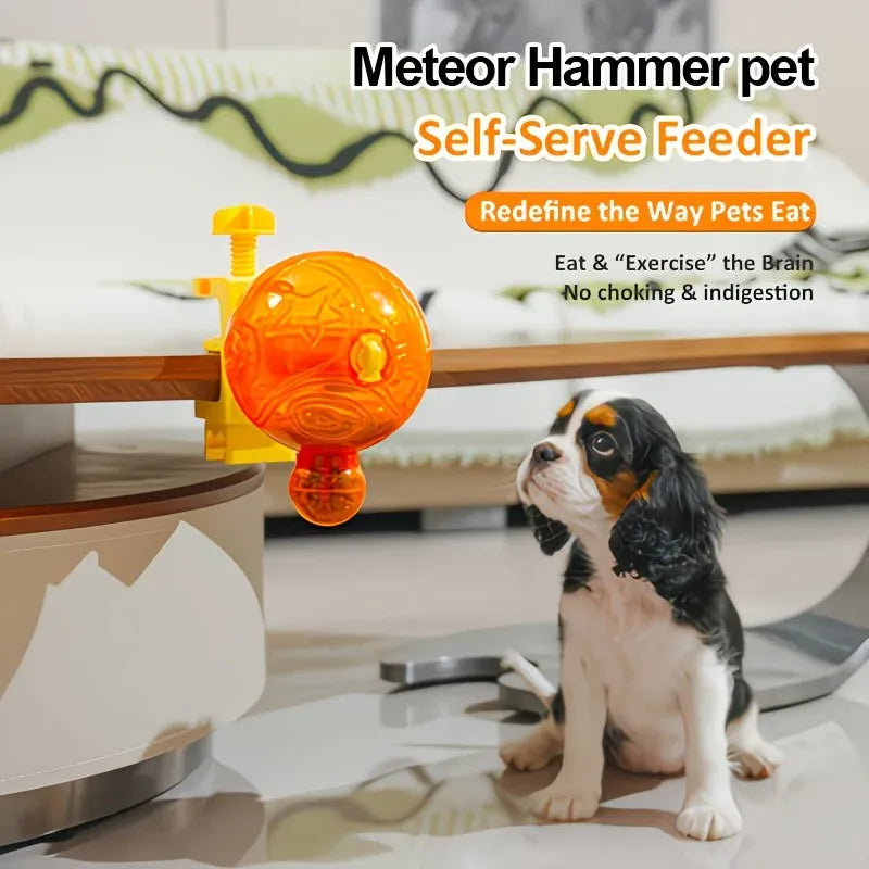 A small Cavalier King Charles Spaniel puppy sits on a glossy floor next to a yellow and orange clip-on ball feeder mounted on a wooden table. Text overlays at the top read 'Meteor Hammer pet Self-Serve Feeder' with additional marketing copy.