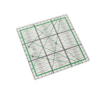 A square quilting ruler with green grid lines and black measurement markings, shown at an angle on a plain white background.