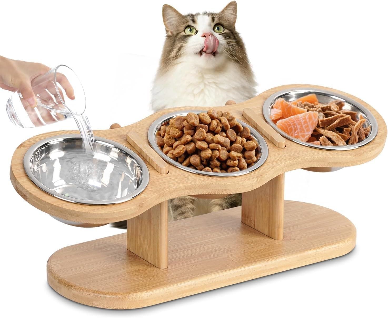  wooden pet feeding station with three stainless steel bowls, shown with kibble and wet food in the center and right bowls, water being poured into the left bowl by a visible human hand, and a cat peeking behind the product on a white backgroun