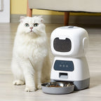 A fluffy white cat sits beside a white and gray automatic pet feeder with a metal food bowl on a light wood floor in a home setting.
