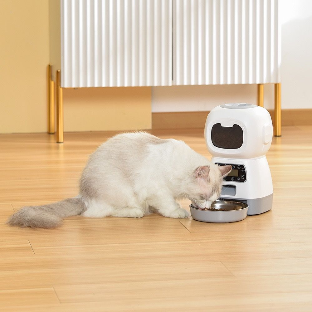 A fluffy white and gray cat is eating from a smart pet feeder on a light wood floor in a modern room.