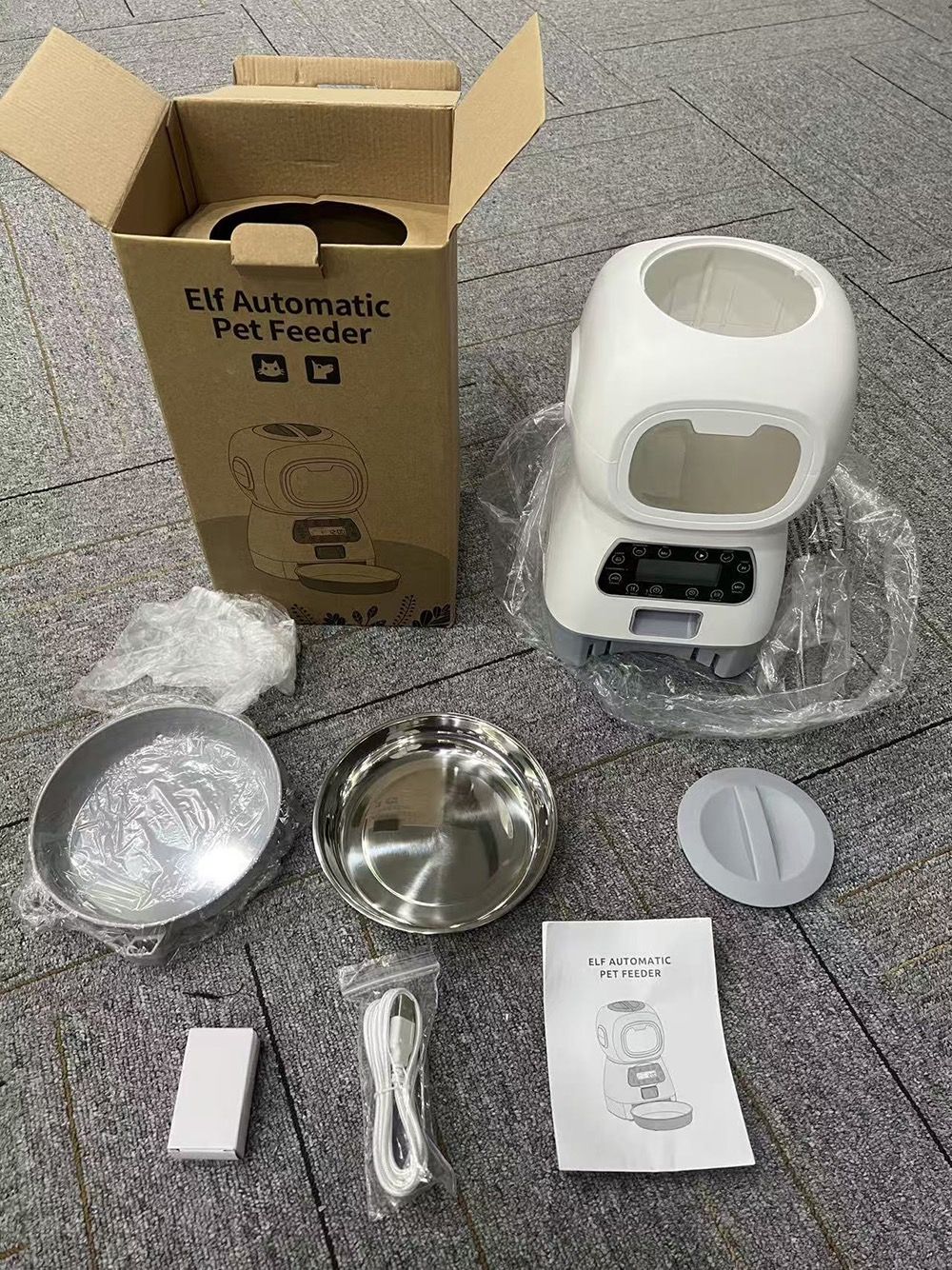 An unboxed pet feeder set on a gray tiled floor, including the white electric feeder, cardboard packaging box, stainless steel bowls, lid, cable, battery, and instruction manual.