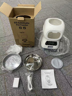 An unboxed pet feeder set on a gray tiled floor, including the white electric feeder, cardboard packaging box, stainless steel bowls, lid, cable, battery, and instruction manual.