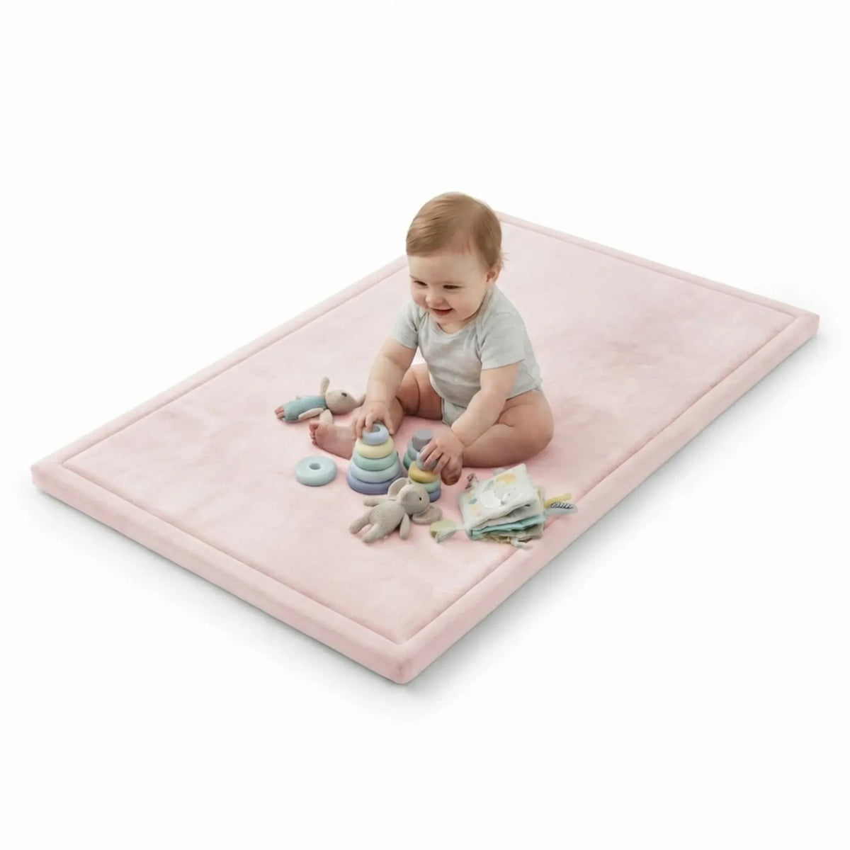 A baby sits on a rectangular pink play mat on a white background, surrounded by small toys.