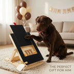 A chocolate brown Labrador retriever sits on a cream rug in a living room, pawing at a black gift box mounted on a wooden stand. The box has a window showing dog treats inside and a small ribbon bow.