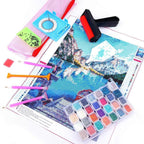 Diamond painting kit with a printed mountain landscape canvas, multicolor beads in a clear plastic tray, paint sticks, a mesh bag, a squeegee, and a roller tool on a white background.