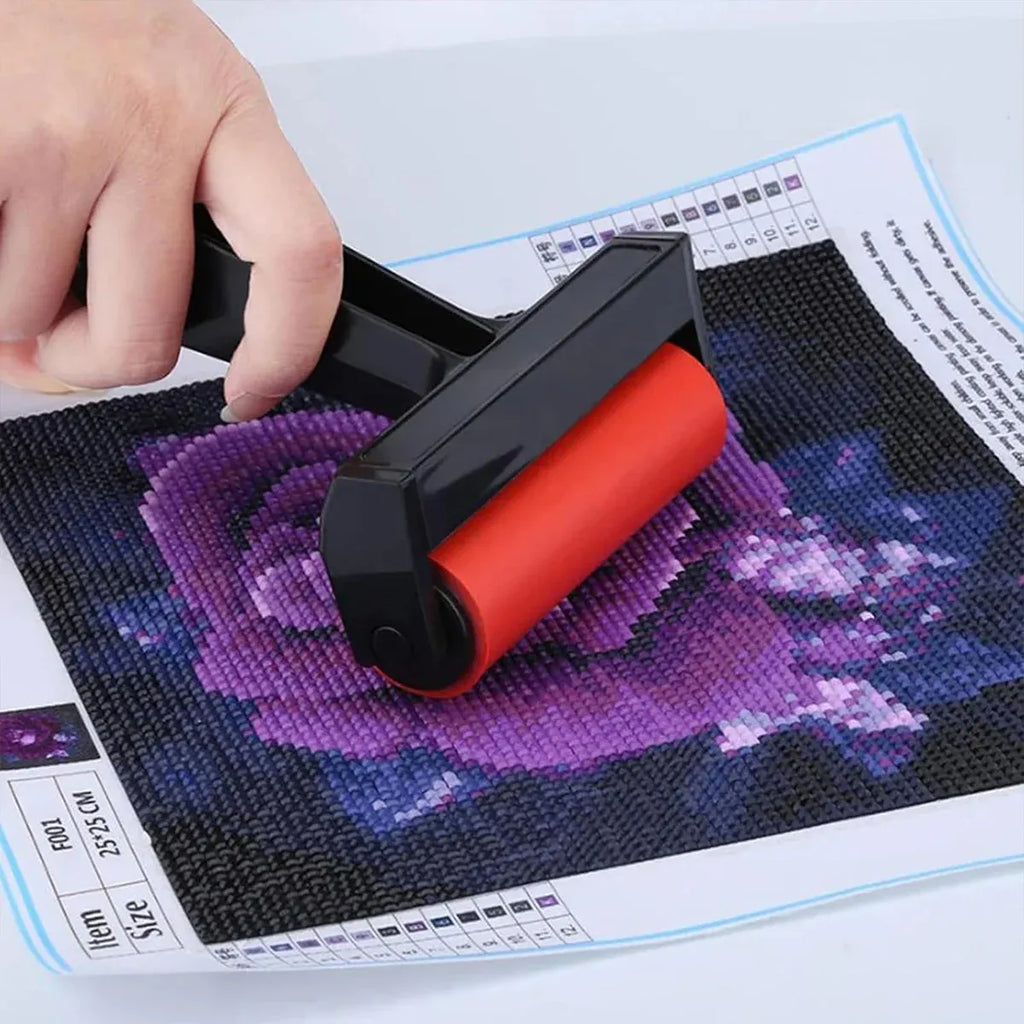 A hand holds a black-and-red diamond painting roller over a printed floral design sheet, showing the tool in use on a craft canvas.