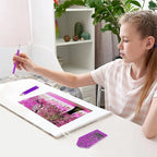 A girl sits at a white desk, holding a purple diamond painting pen over a colorful canvas showing a pink floral scene. A matching purple diamond painting kit piece is placed on the desk, with a pencil nearby.
