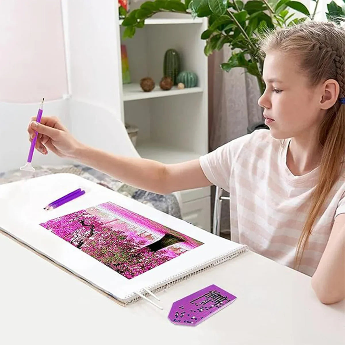 A girl sits at a white desk, holding a purple diamond painting pen over a colorful canvas showing a pink floral scene. A matching purple diamond painting kit piece is placed on the desk, with a pencil nearby.