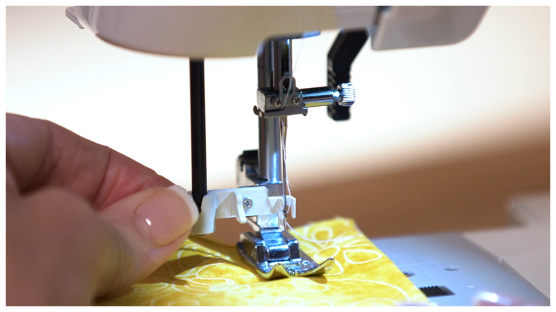 Close-up of a sewing machine needle on yellow fabric with a hand guiding it.