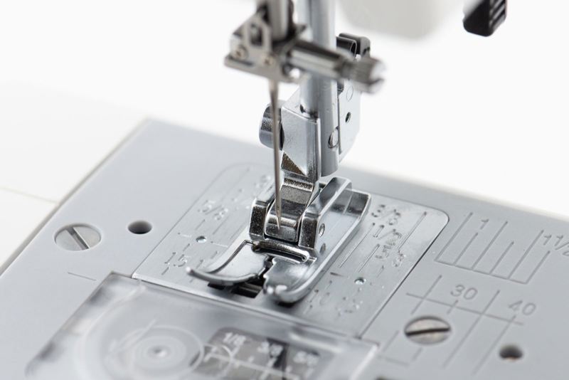 Close-up of a sewing machine needle and presser foot assembly on a light gray sewing machine bed with measurement markings.