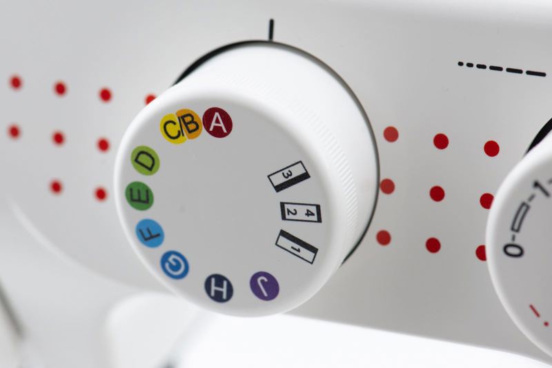 Close-up of a white sewing machine dial with colorful thread color indicators and stitch number markings, set against a white machine body with red dot accents.