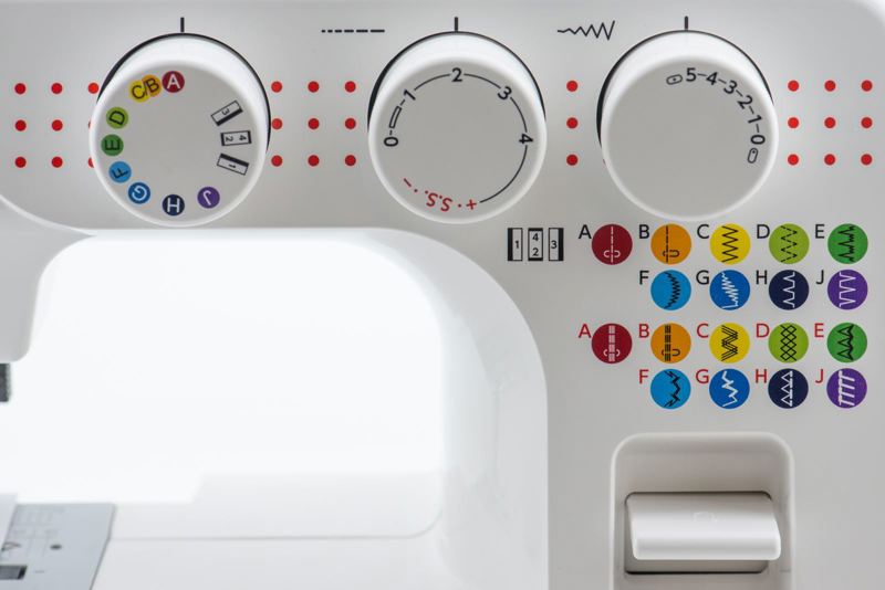 Close-up of a white sewing machine head showing three control dials, red dotted guide markings, and a grid of colorful stitch pattern icons labeled A through J.

Details