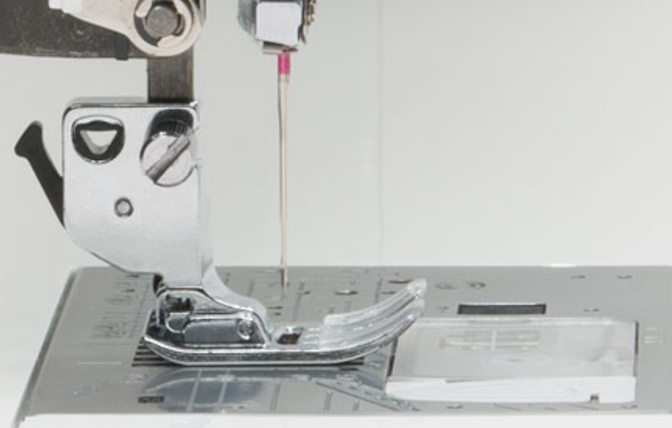 Close-up of a sewing machine presser foot and needle area, showing a clear plastic foot cover and a silver metal presser assembly on a white sewing machine bed.
