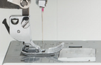 Close-up of a sewing machine presser foot and needle area, showing a clear plastic foot cover and a silver metal presser assembly on a white sewing machine bed.