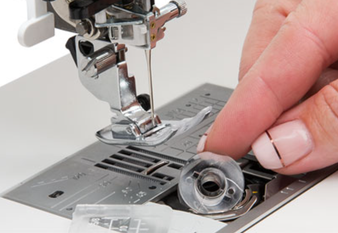 Close-up of a hand placing a clear plastic bobbin onto a sewing machine feed dog area.