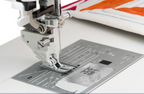 Close-up of a Single needles Acufeed  foot in the sewing machine needle area and metal plate with a clear cover, on a white machine bed. A partially finished quilt with orange and pink stitching is visible in the background.