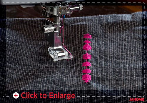 A close-up of a sewing machine needle and presser foot stitching dark gray fabric, with a vertical row of bright pink satin stitch embellishments being created. A 'Click to Enlarge' overlay is visible at the bottom.